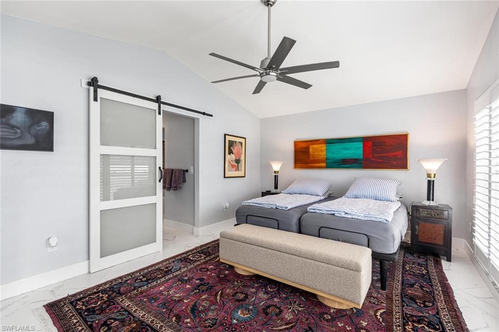 Bedroom showing porcelain tile flooring, a barn door, ceiling fan, and lofted ceiling. Windows have wooden plantation shutters.
