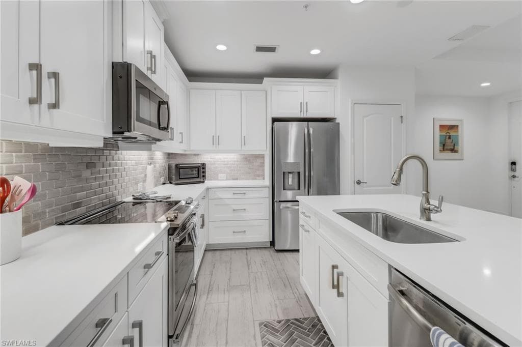 Light, bright kitchen with white cabinetry, stainless steel appliances, decorative backsplash, sink, and under-counter lighting