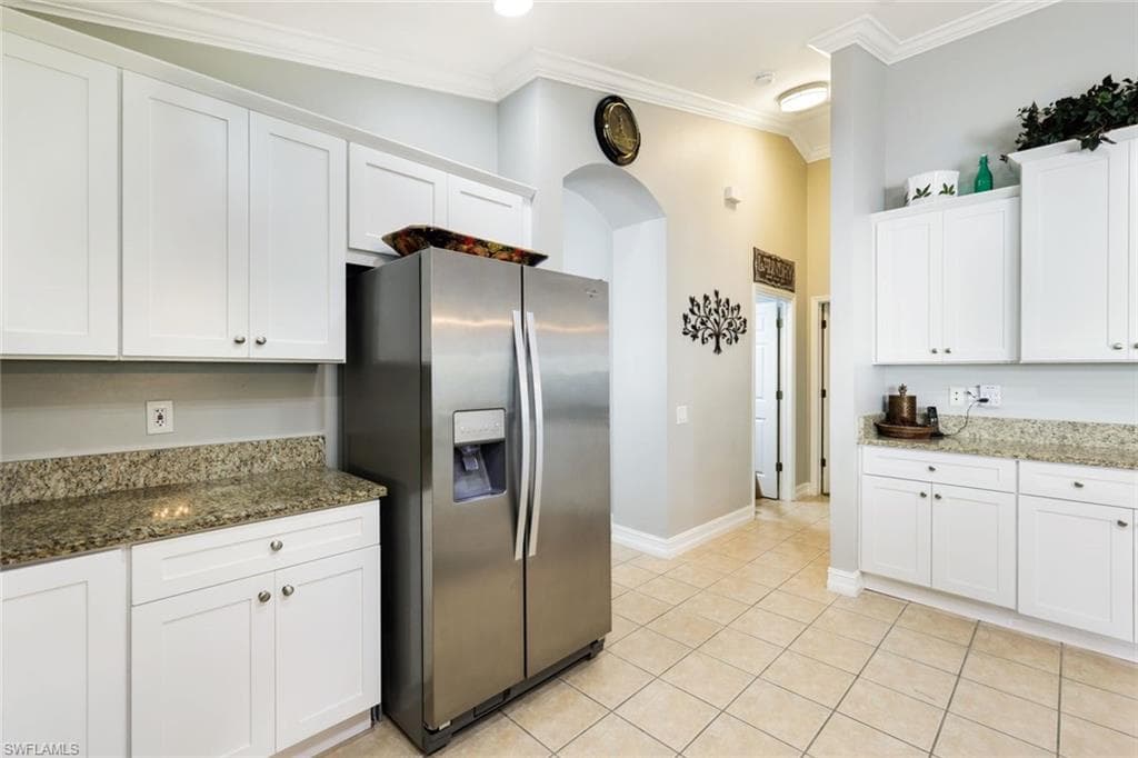 Kitchen with light tile patterned floors, ornamental molding, stone countertops, white cabinets, and stainless steel fridge