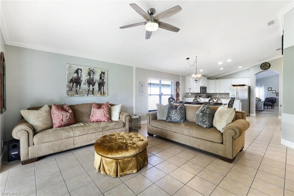 Living area with arched walkways, light tile patterned floors, ornamental molding, vaulted ceiling, and ceiling fan with notable chandelier