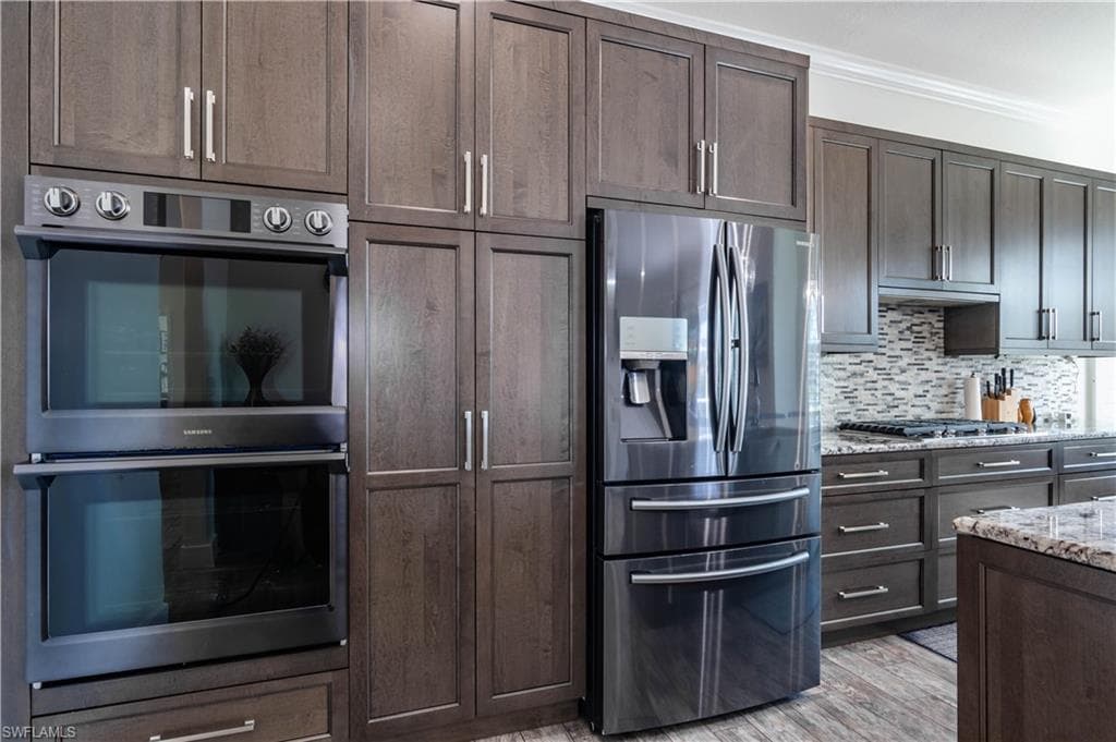 Kitchen with stainless steel appliances, tasteful backsplash, dark brown cabinets, light stone counters, and crown molding