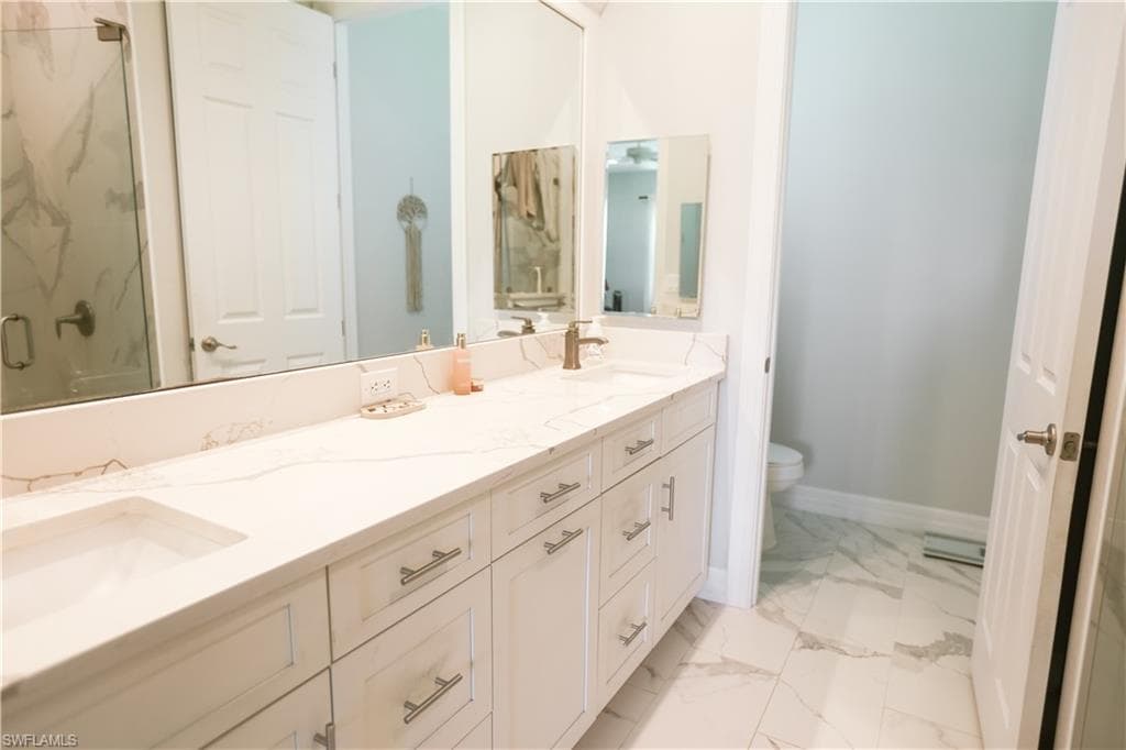 Bathroom featuring double vanity, marble look tiles, toilet, a marble finish shower, and baseboards