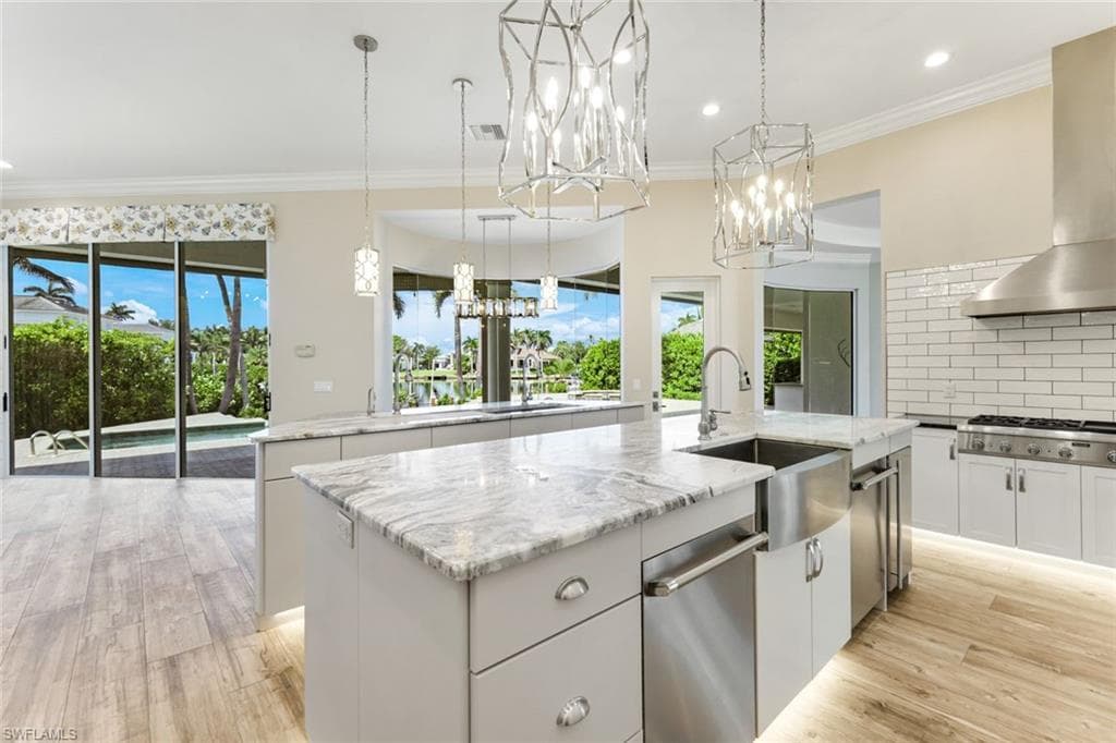Kitchen with a kitchen island with sink, wall chimney range hood, a chandelier, appliances with stainless steel finishes, and white cabinets