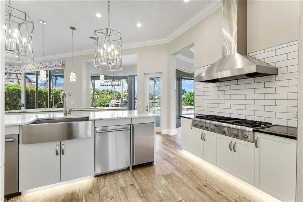 Kitchen featuring appliances with stainless steel finishes, wall chimney range hood, light wood finished floors, a chandelier, and backsplash