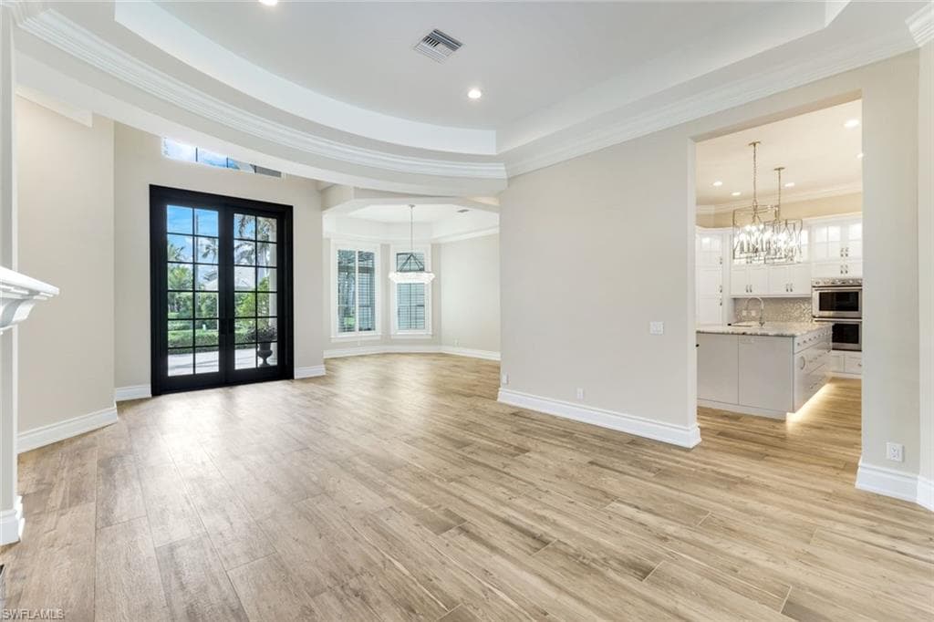 Unfurnished living room with a tray ceiling, a chandelier, light wood finished floors, french doors, and crown molding