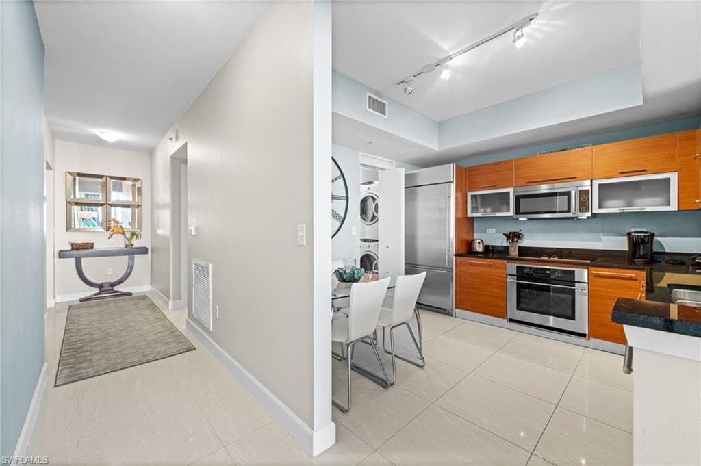 Kitchen with brown cabinetry, appliances with stainless steel finishes, light tile patterned floors, stacked washer / dryer, and rail lighting