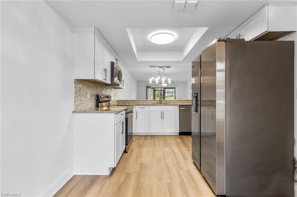 Kitchen with stainless steel appliances, white cabinets, light stone countertops, a raised ceiling, and hanging light fixtures