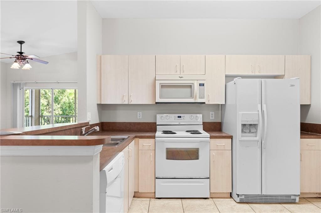Kitchen featuring light brown cabinetry, white appliances, a peninsula, and dark countertops