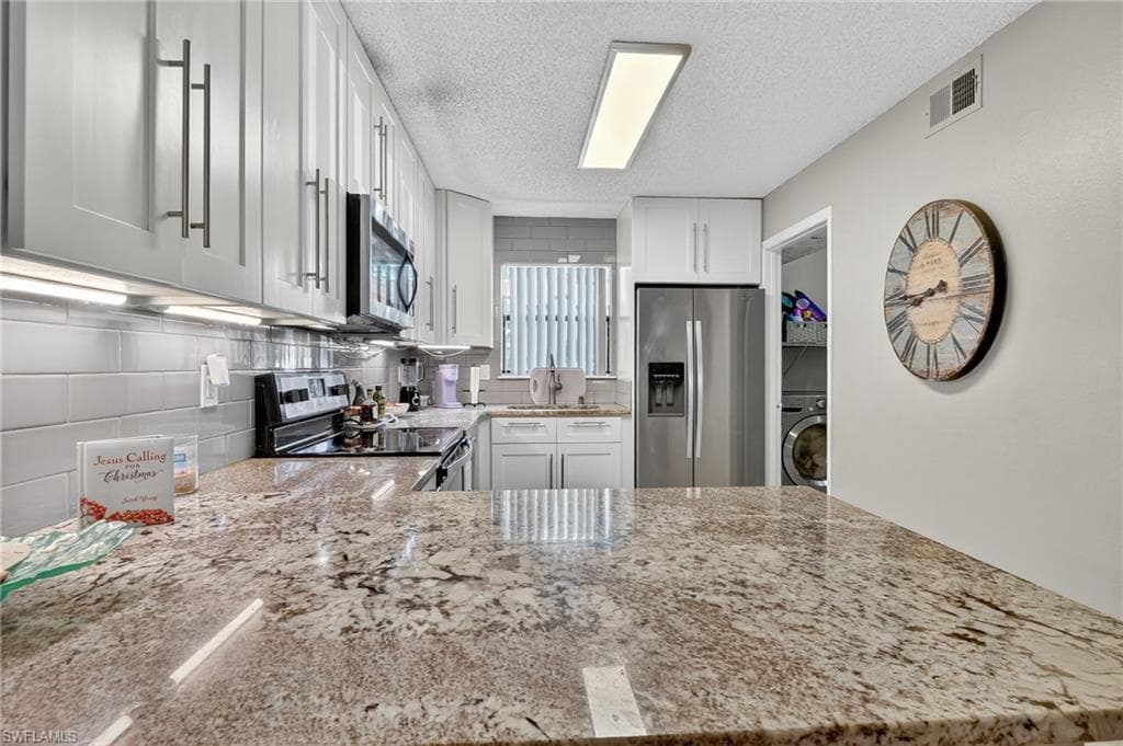 Kitchen featuring stainless steel appliances, light stone counters, white cabinetry, washer / dryer, and a textured ceiling