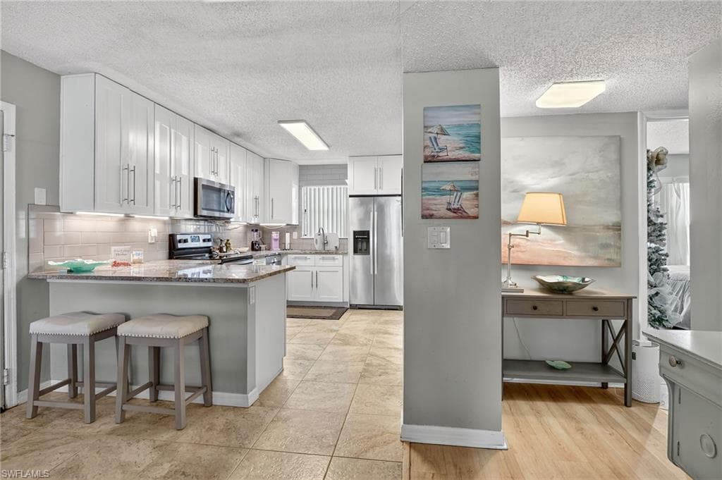Kitchen with a textured ceiling, stainless steel appliances, light stone counters, white cabinetry, and a kitchen bar