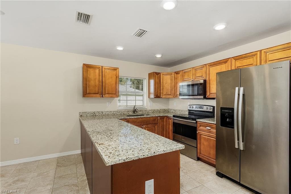 Kitchen with appliances with stainless steel finishes, a peninsula, light stone counters, brown cabinetry, and recessed lighting