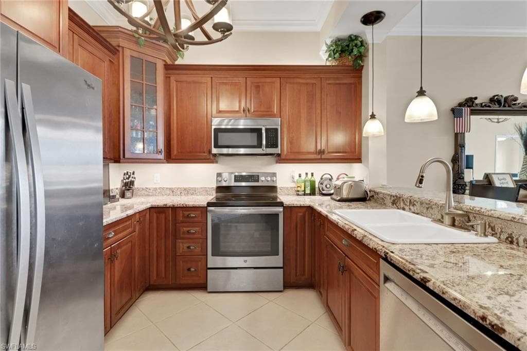 Kitchen featuring brown cabinetry, appliances with stainless steel finishes, hanging light fixtures, ornamental molding, and light stone countertops