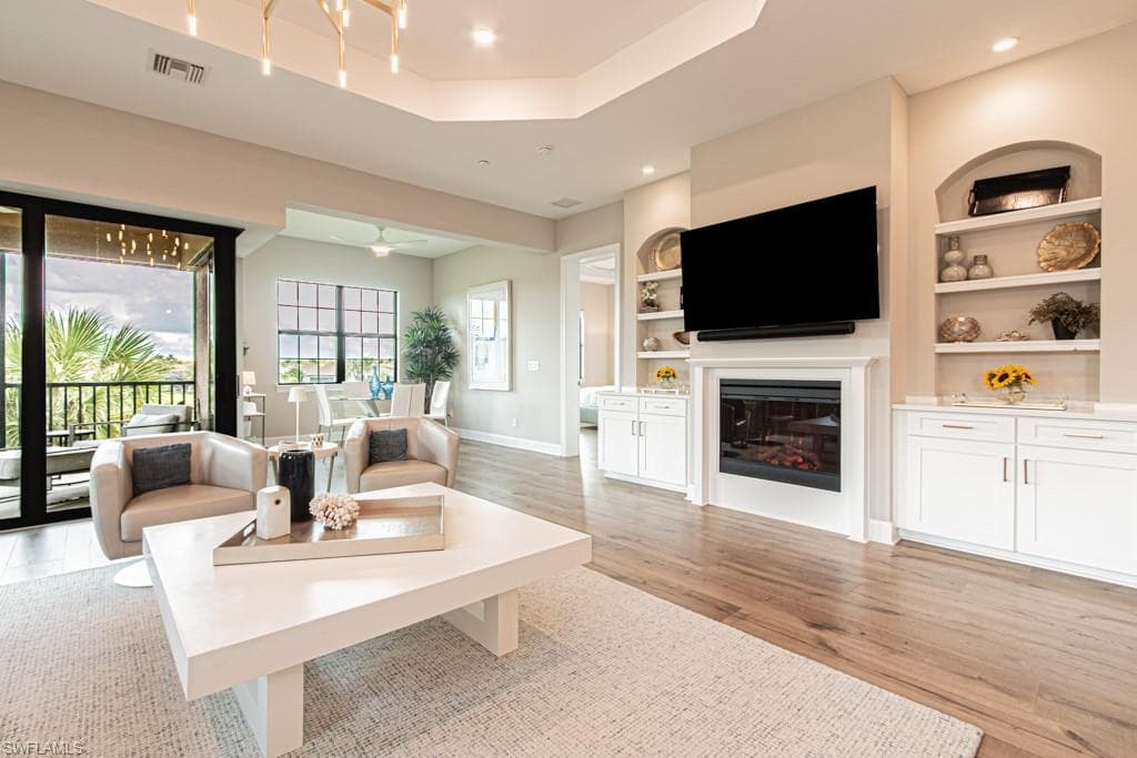 Living room featuring a glass covered fireplace, light wood-type flooring, a raised ceiling, recessed lighting, and built in features