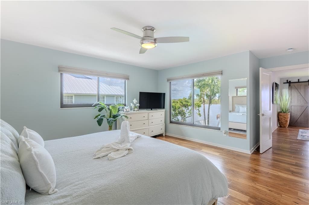 Bedroom featuring a barn door, multiple windows, light wood-style flooring, and a ceiling fan - Virtually Edited Image