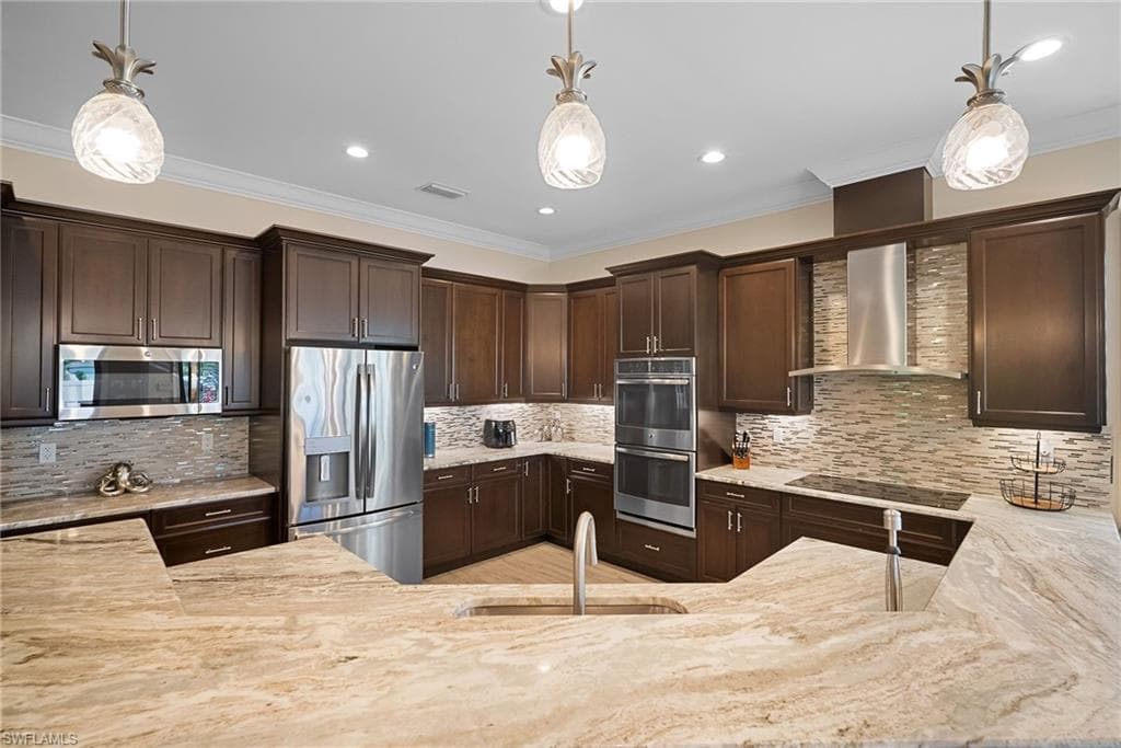Kitchen with dark brown cabinetry, ornamental molding, stainless steel appliances, decorative backsplash, and wall chimney exhaust hood