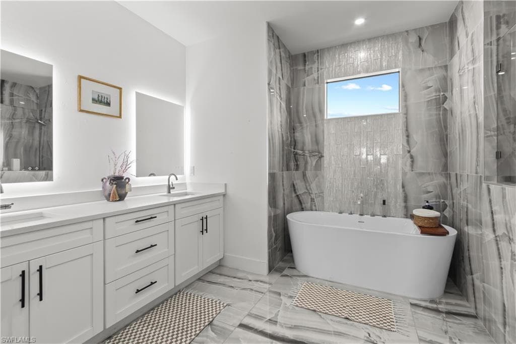 Bathroom with a marble finish shower, double vanity, light marble finish flooring, a freestanding tub, and tile walls