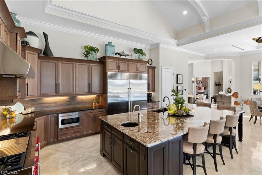 Kitchen featuring crown molding, dark wood finish cabinets, dark stone countertops, built in appliances, and a kitchen island with sink
