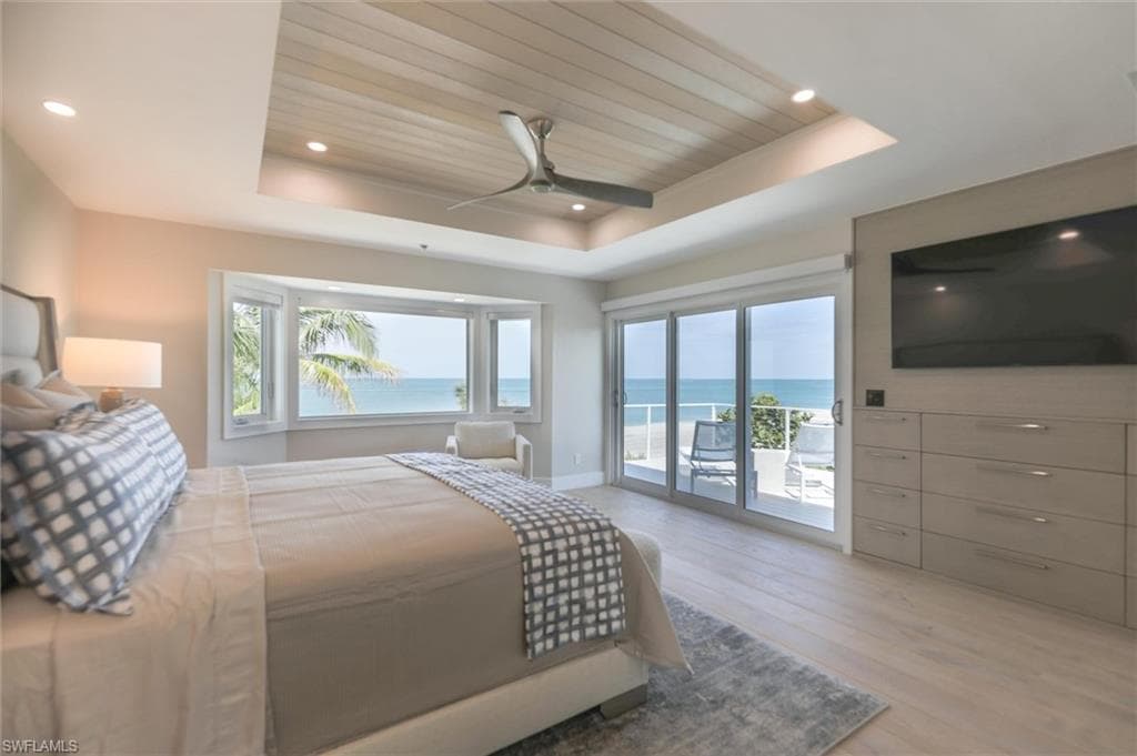 Bedroom with light wood-type flooring, a water view, access to exterior, ceiling fan, and a raised ceiling