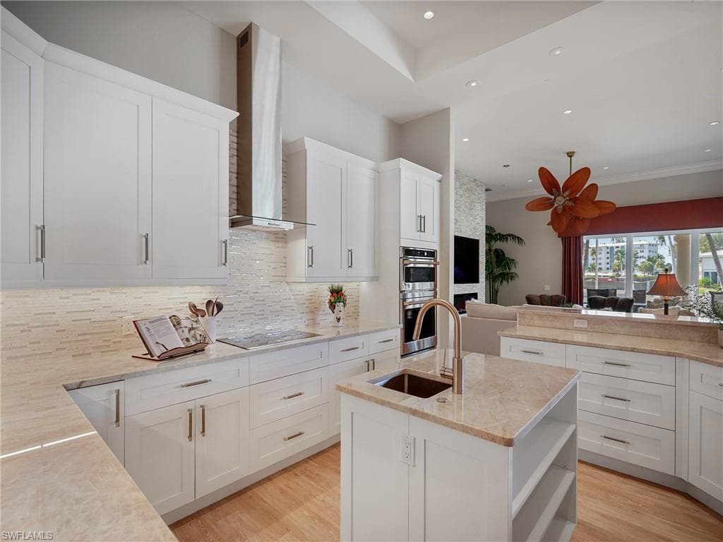 Kitchen featuring open shelves, a center island with sink, decorative backsplash, wall chimney range hood, and light stone countertops