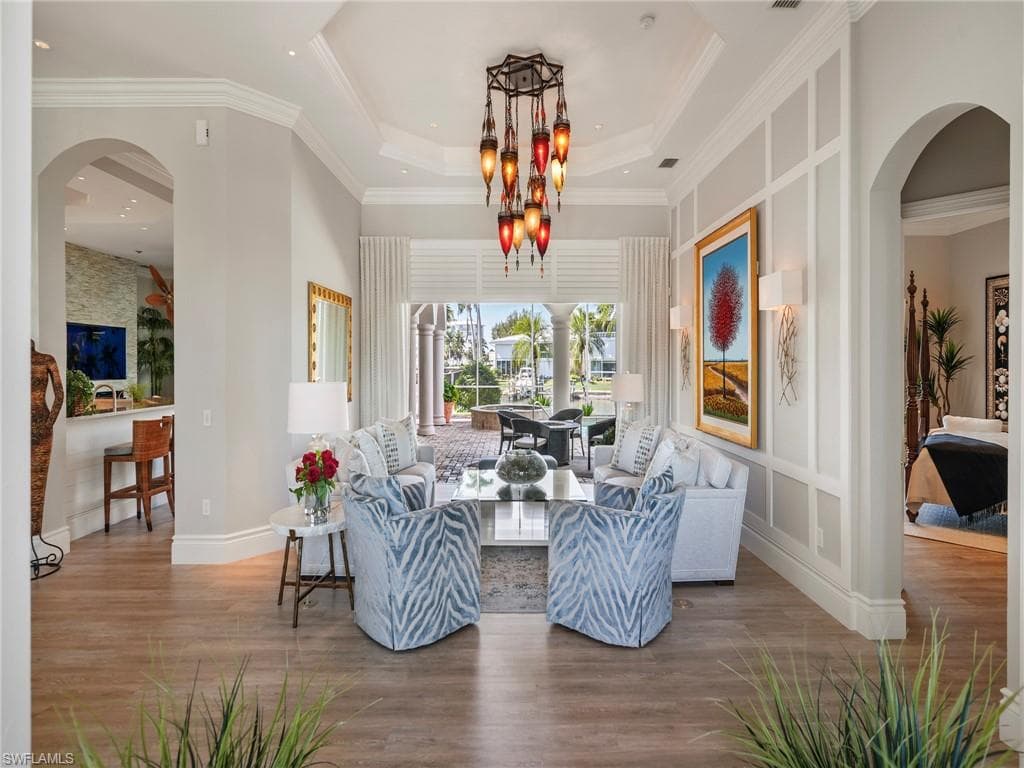Living area with arched walkways, wood finished floors, ornamental molding, a tray ceiling, and a chandelier