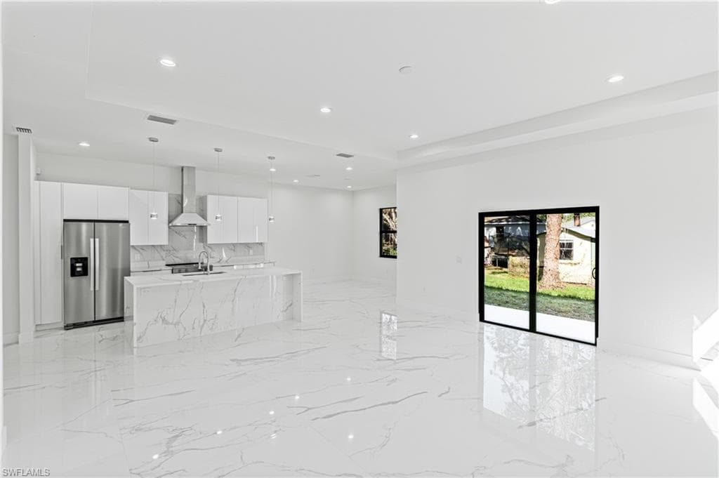 Kitchen featuring stainless steel refrigerator with ice dispenser, white cabinets, wall chimney exhaust hood, light stone counters, and modern cabinets