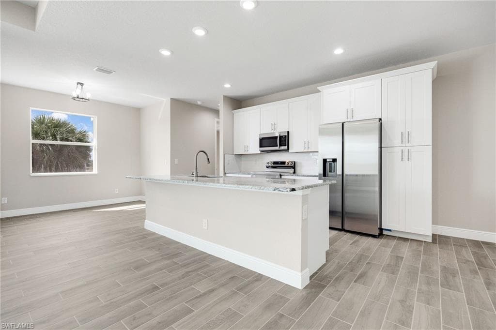 Kitchen with appliances with stainless steel finishes, light stone counters, white cabinets, an island with sink, and wood tiled floors-Finishes may vary