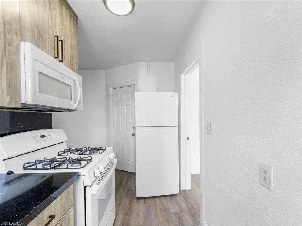 Kitchen with white appliances, light wood-style flooring, a textured wall, and dark stone counters