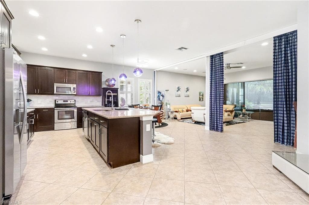 Kitchen featuring appliances with stainless steel finishes, pendant lighting, open floor plan, light stone counters, and dark brown cabinets