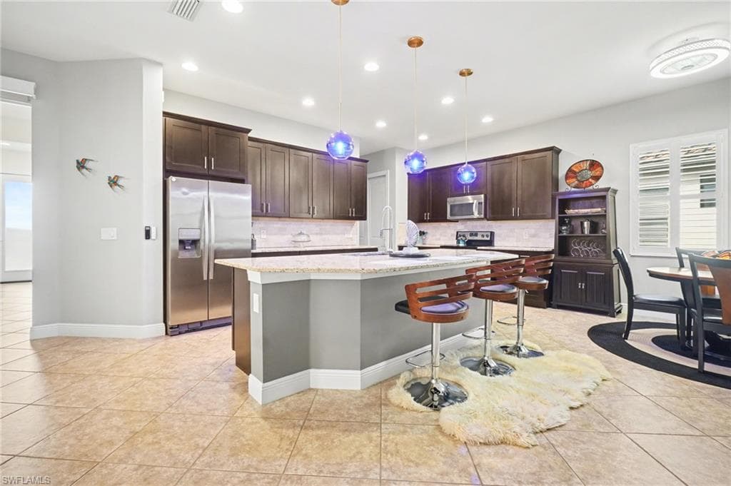 Kitchen featuring dark brown cabinets, appliances with stainless steel finishes, a breakfast bar area, light stone countertops, and decorative light fixtures