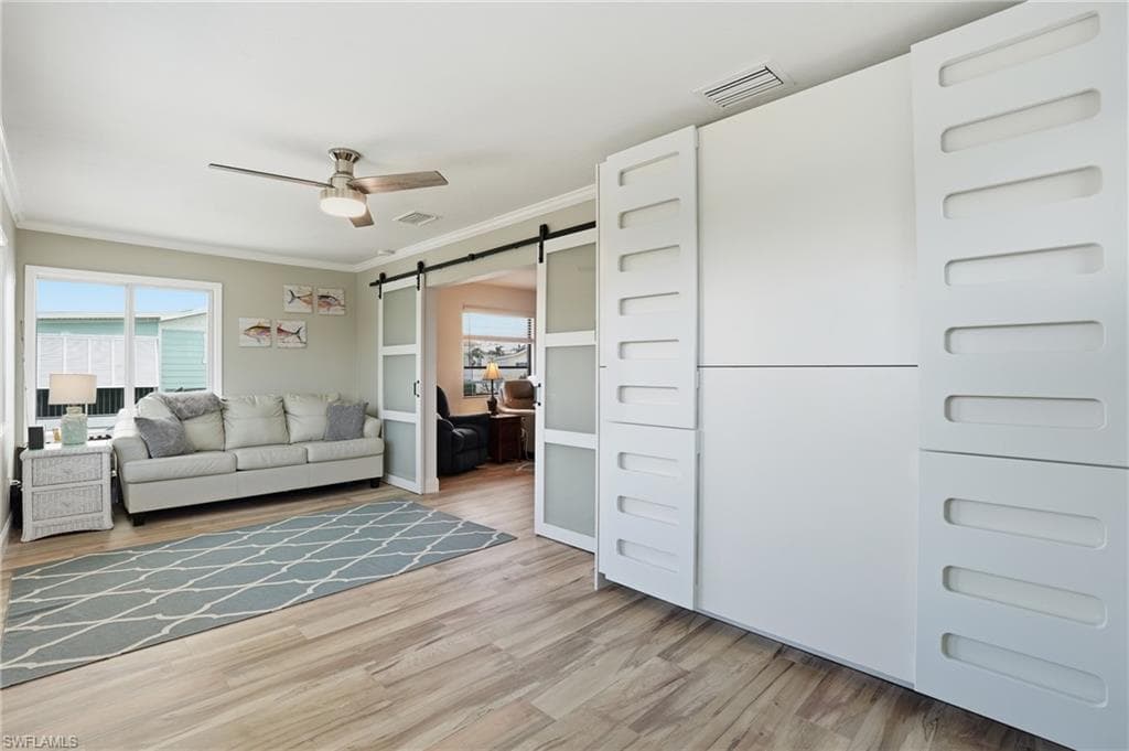 Den featuring a barn door, crown molding, ceiling fan, plenty of natural light, and light wood-style floors - Virtually Edited Image