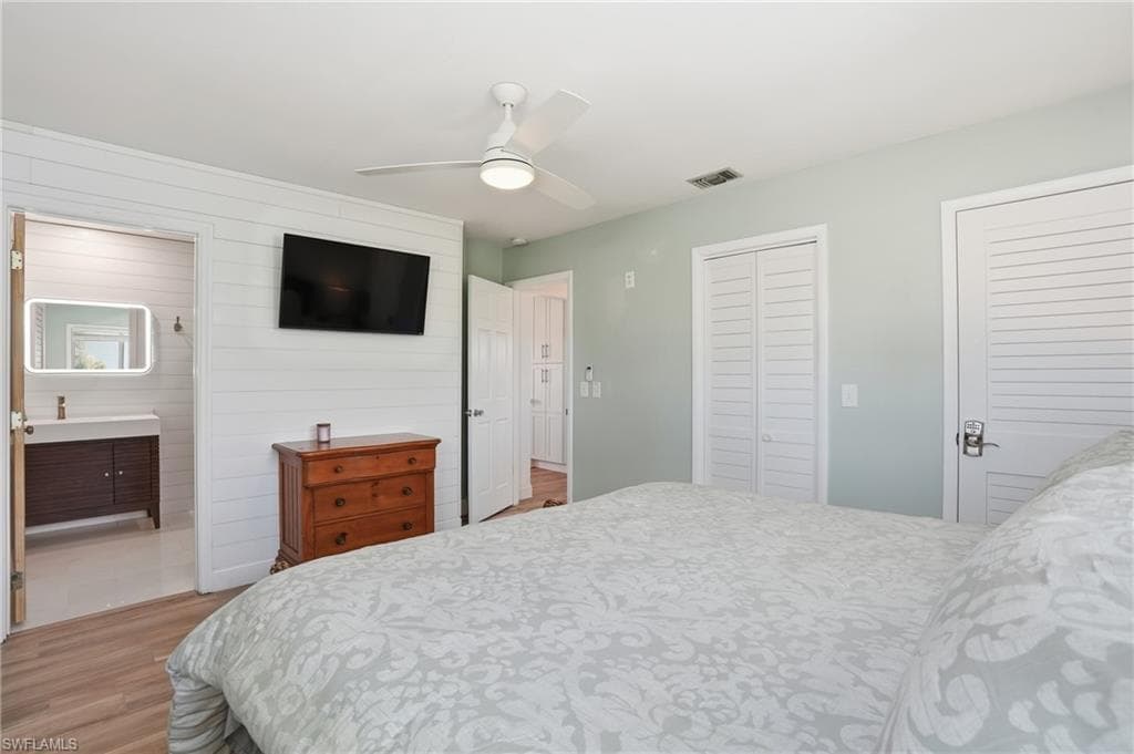 Bedroom featuring light wood-style floors, two closets, ceiling fan, wooden walls, and ensuite bath - Virtually Edited Image