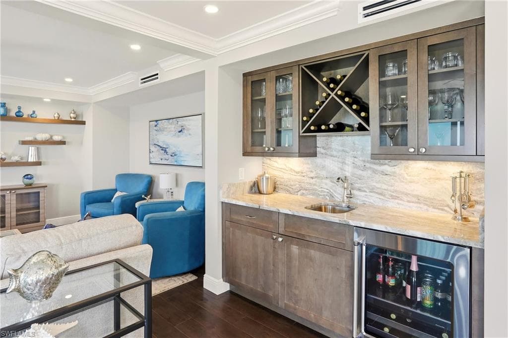 Indoor wet bar with beverage cooler, backsplash, light stone countertops, ornamental molding, and dark wood-style floors