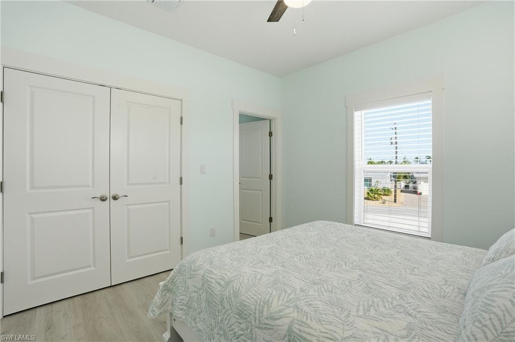 Bedroom featuring light blue walls, wood-finish flooring, a window with blinds, and white bi-fold closet doors - Virtually Edited Image