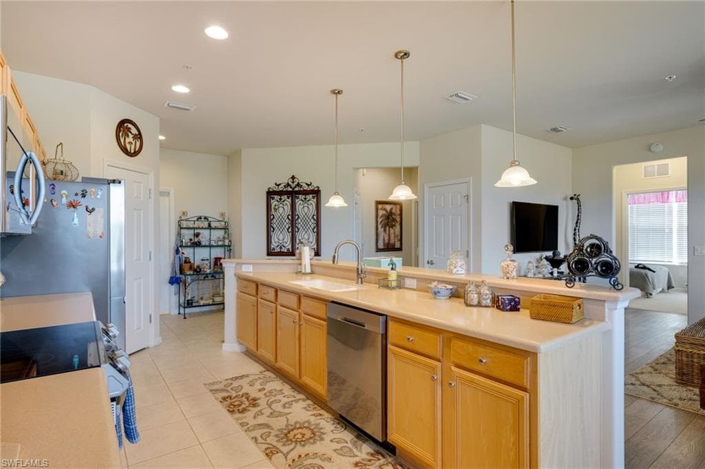 Kitchen featuring light countertops, appliances with stainless steel finishes, pendant lighting, a kitchen island with sink, and light brown cabinetry