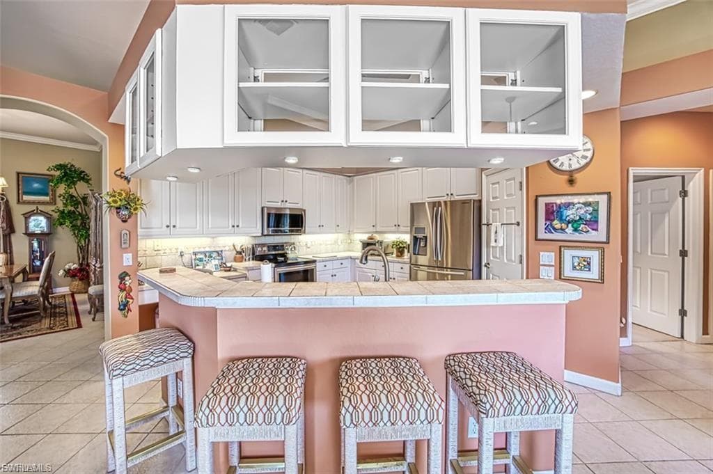 Kitchen with light tile patterned flooring, glass insert cabinets, a breakfast bar area, stainless steel appliances, and ornamental molding