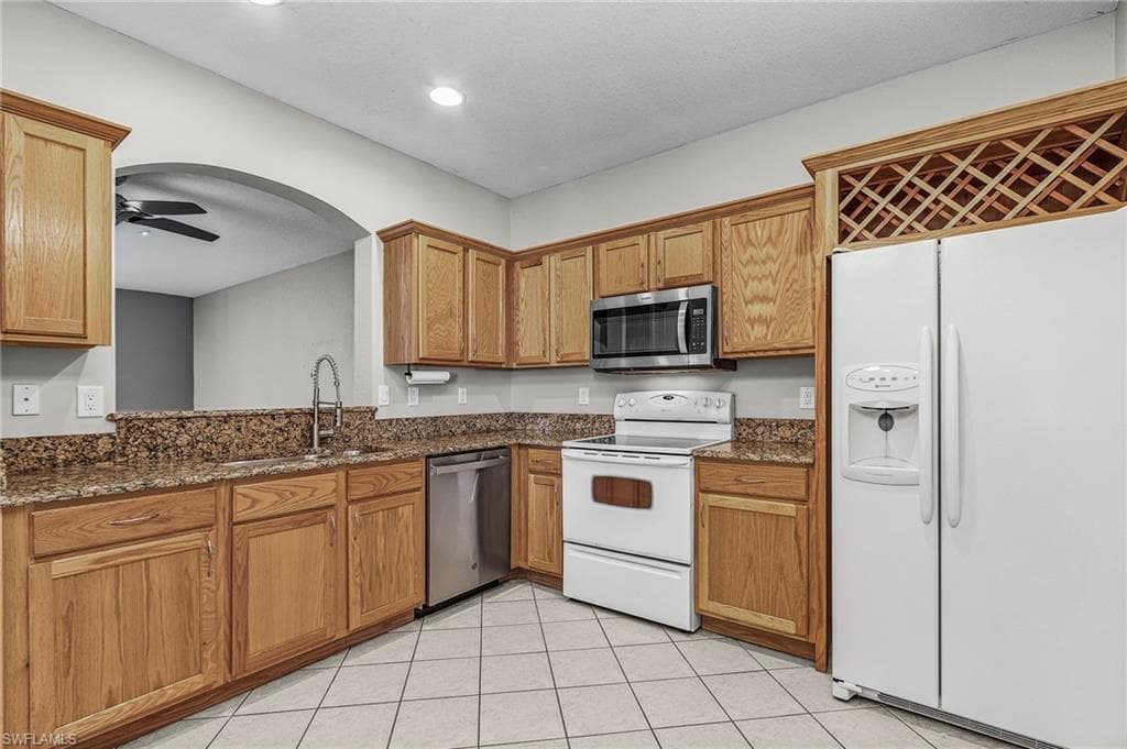 Kitchen featuring stainless steel appliances, dark stone countertops, ceiling fan, arched walkways, and brown cabinetry