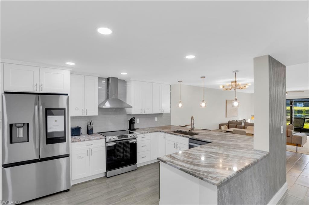 Kitchen with appliances with stainless steel finishes, a peninsula, hanging light fixtures, wall chimney range hood, and white cabinets
