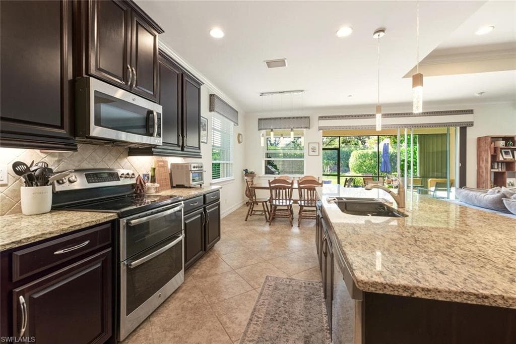 Kitchen with stainless steel appliances, crown molding, dark brown cabinets, light stone countertops, and pendant lighting
