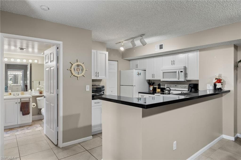 Kitchen with a peninsula, dark countertops, white cabinets, white appliances, and a textured ceiling