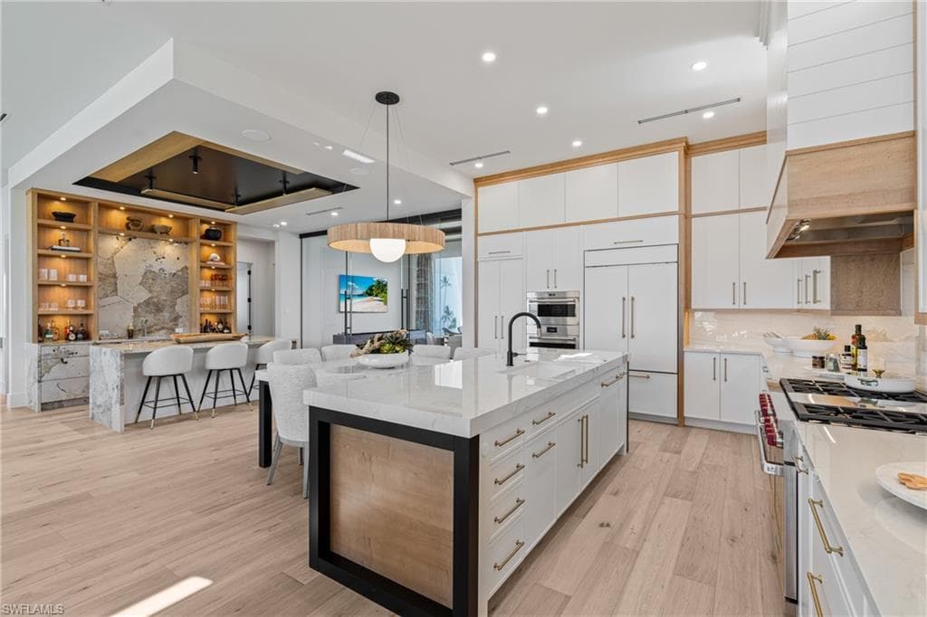 Kitchen with light stone counters, high quality appliances, a large island with sink, hanging light fixtures, and recessed lighting