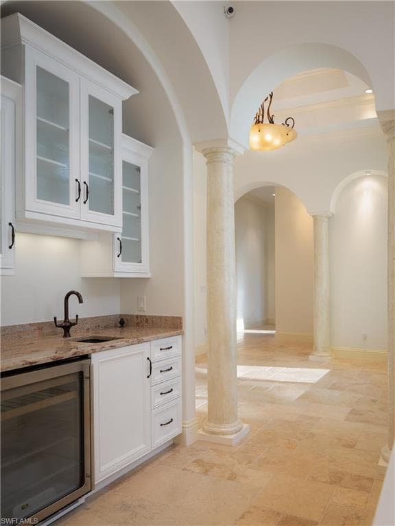 Bar with wine cooler, white cabinetry, light stone countertops, glass insert cabinets, and arched walkways
