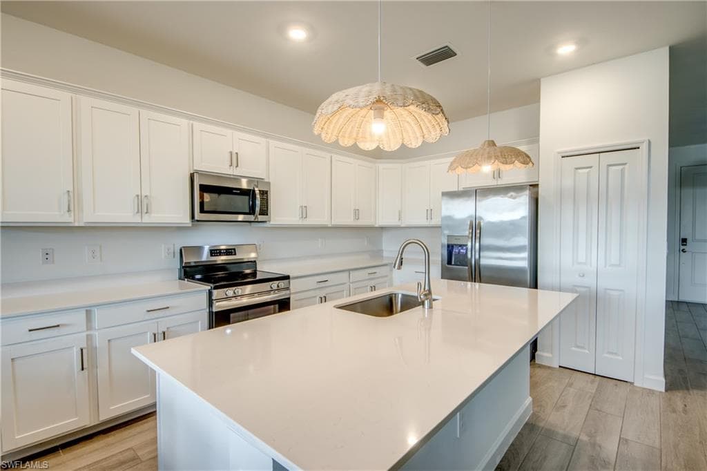 Kitchen featuring appliances with stainless steel finishes, white cabinets, light wood-style floors, a center island with sink, and hanging light fixtures