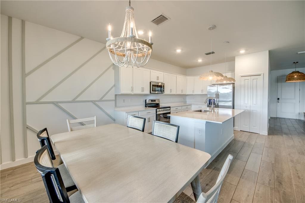 Kitchen featuring white cabinets, a kitchen island with sink, appliances with stainless steel finishes, light wood finished floors, and decorative light fixtures