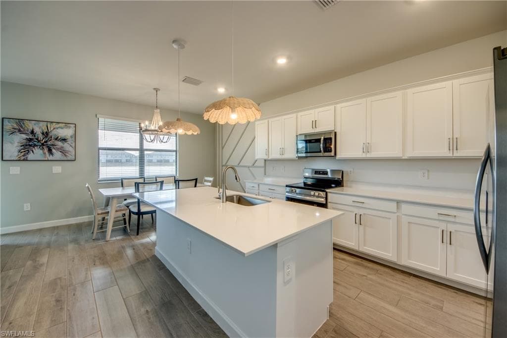 Kitchen with white cabinets, appliances with stainless steel finishes, light wood-style floors, pendant lighting, and an island with sink