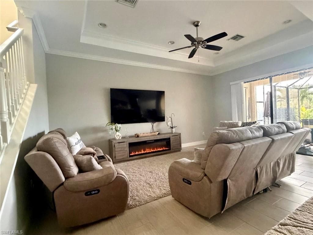 Living room with wood finished floors, a glass covered fireplace, ceiling fan, a tray ceiling, and ornamental molding
