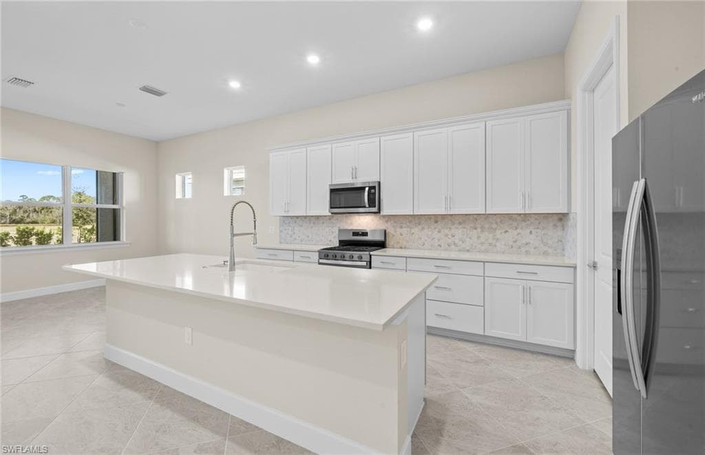 Kitchen featuring backsplash, stainless steel appliances, white cabinets, a center island with sink, and light stone countertops