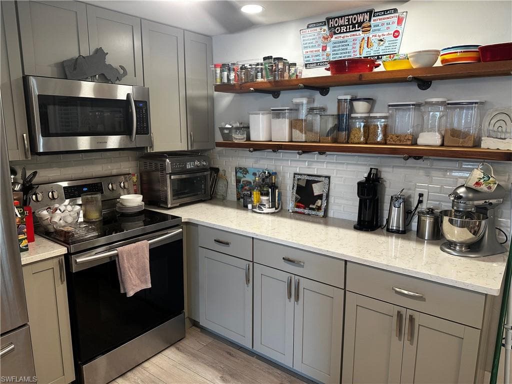 Kitchen with gray cabinets, open shelves, stainless steel appliances, and light stone countertops