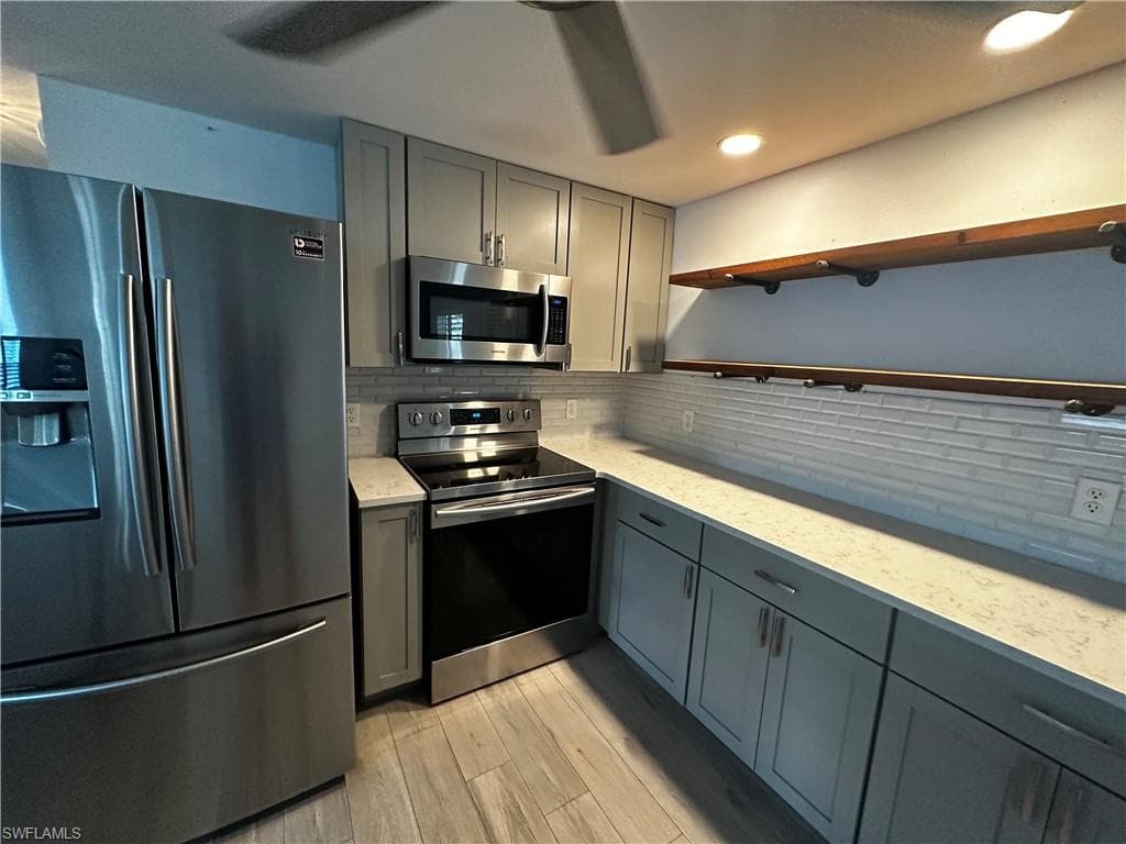 Kitchen with stainless steel appliances, gray cabinetry, open shelves, light wood-style floors, and light stone countertops