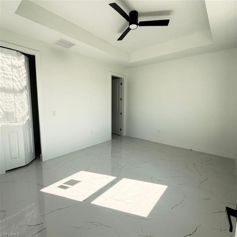 Empty room with a raised ceiling, ceiling fan, and light marble finish flooring