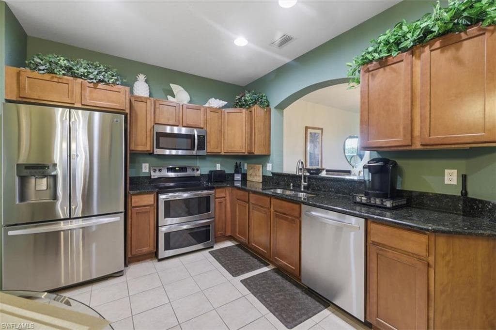 Kitchen featuring stainless steel appliances, dark stone countertops, wood finish cabinets, light tile patterned floors, and arched walkways